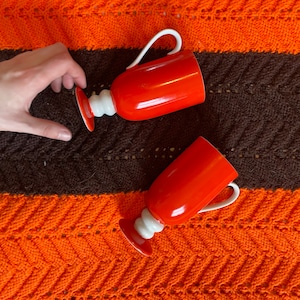 Puede incluir: Dos tazas de cerámica roja con detalles blancos. Las tazas tienen una forma redondeada y un asa en un lado. Las tazas están sobre una manta de punto marrón y naranja.
