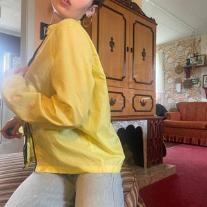 May include: A yellow lightweight jacket with a zipper closure. The jacket is worn by a person with a light brown hair and a gold hoop earring.
