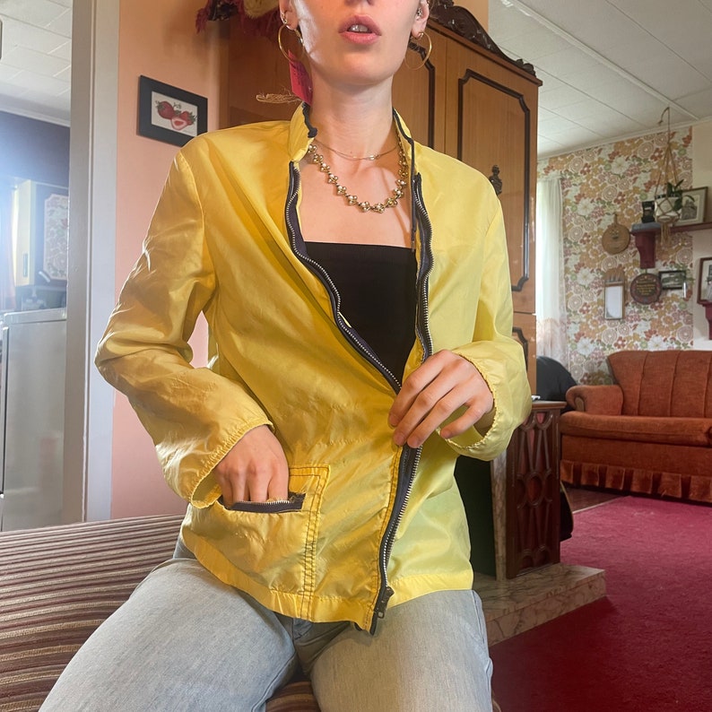 May include: A yellow zippered jacket with a black tank top underneath. The jacket has a zipper closure and two pockets. The person is wearing gold hoop earrings and a gold necklace.