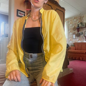 May include: A yellow lightweight jacket with a zipper closure. The jacket has a black zipper and a black collar. The model is wearing a black tank top and blue jeans.