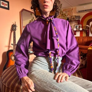 May include: A vibrant purple blouse with long sleeves and a patterned necktie. The blouse has a classic design with a high collar and buttoned cuffs. The necktie features orange and purple patterns. The image is set in a retro-style room with a guitar and a dog.