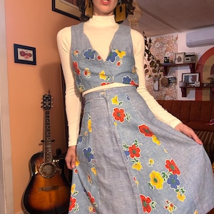 May include: A light blue denim two-piece outfit with a floral pattern in red, yellow, and blue. The set includes a cropped vest and a midi skirt. The outfit is worn with a white turtleneck.