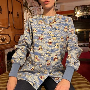 May include: A blue long-sleeved shirt with a white background and a pattern of brown bears wearing winter hats and scarves. The bears are skiing, ice skating, and sledding. The shirt has a button-up front and a collar.