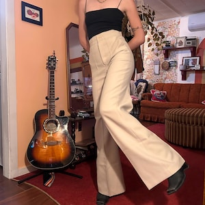Puede incluir: Pantalones acampanados color crema de cintura alta con cierre de botón. Los pantalones se combinan con un top negro y botas negras. Una guitarra acústica es visible en el fondo.