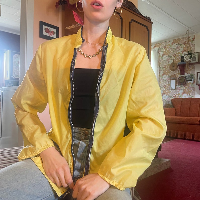 May include: A yellow lightweight jacket with a zipper closure and a black tank top underneath. The jacket has a stand-up collar and long sleeves.