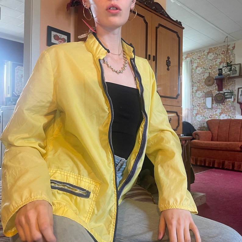 May include: A yellow lightweight jacket with a zipper closure and a zippered pocket on the left side. The jacket has a black zipper and a silver chain necklace.