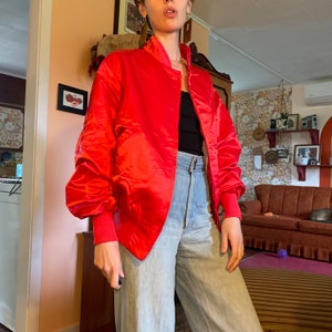 May include: A bright red satin bomber jacket with a zipper closure. The jacket has a stand-up collar and long sleeves.