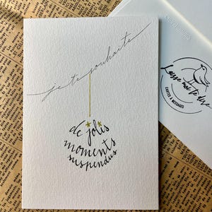 Carte de vœux blanche avec écriture noire à la plume.
Le message dit :" Je te souhaite de jolis moments suspendus".
Des étoiles dorées remplacent le point sur les i. Le texte forme une boule de Noël.