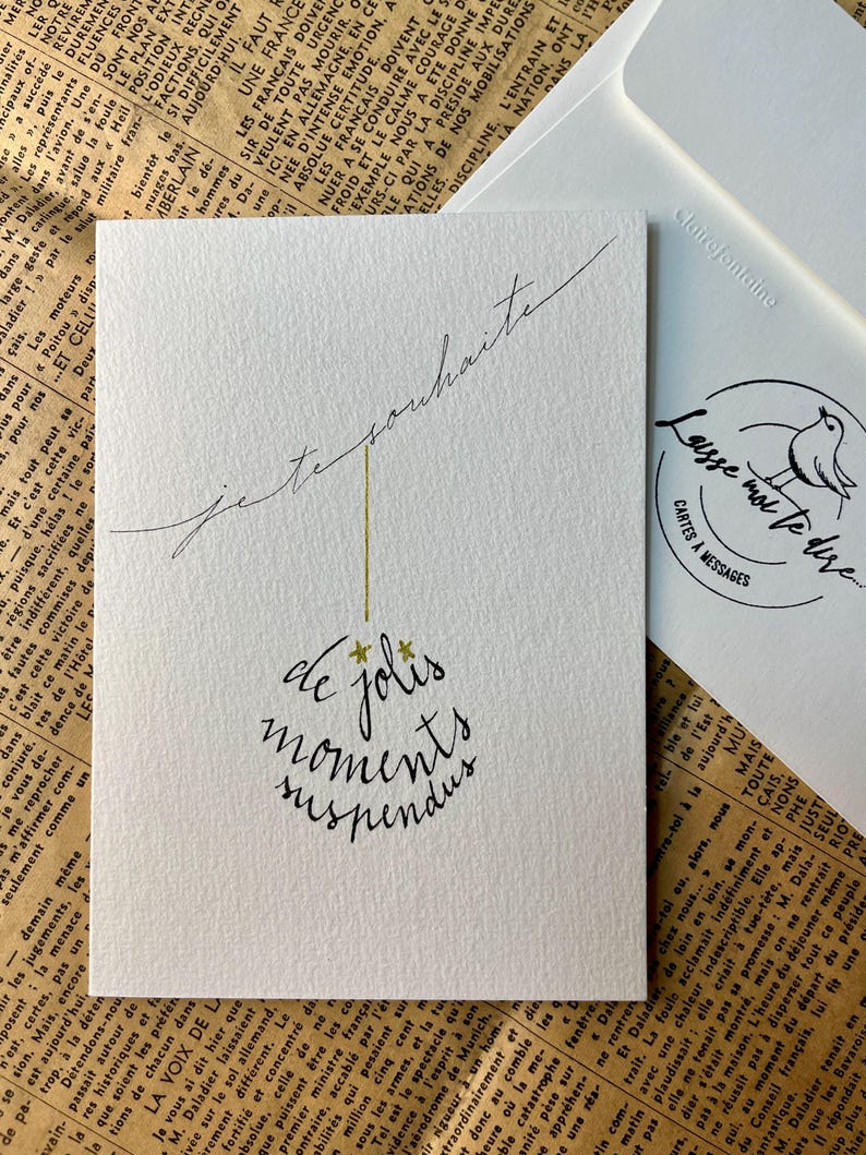 Carte de vœux blanche avec écriture noire à la plume.
Le message dit :" Je te souhaite de jolis moments suspendus".
Des étoiles dorées remplacent le point sur les i. Le texte forme une boule de Noël.