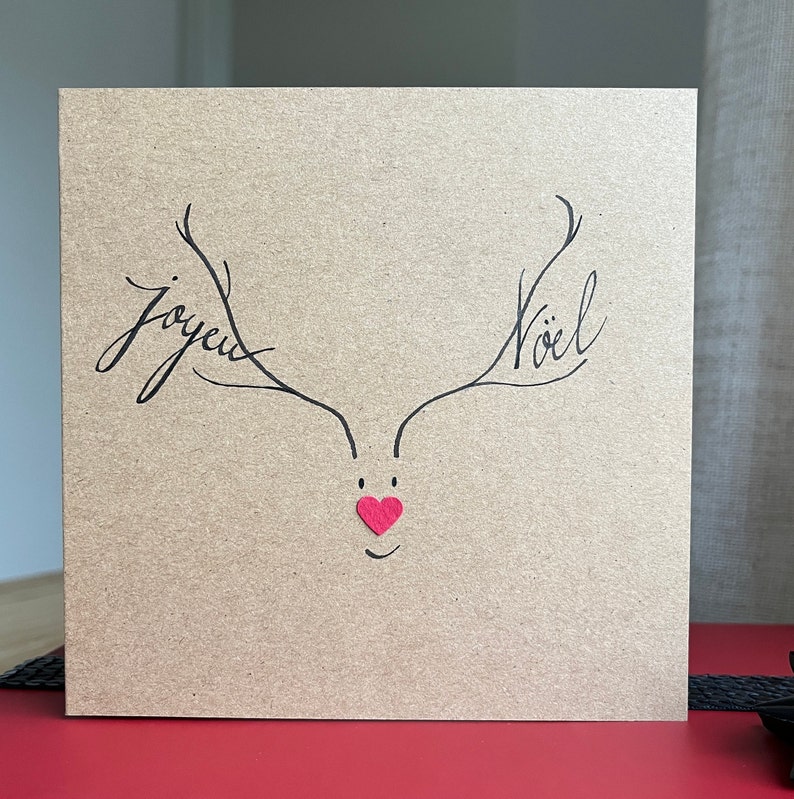 Carte double de Noël en carton couleur kraft.Les bois d'un cerf écrivent Joyeux Noël, il sourit, son nez est un cœur rouge.