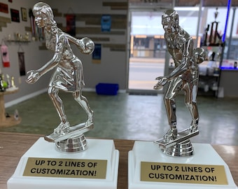 May include: Two silver bowling trophies on white bases with gold plates. The trophies feature a bowler in action, holding a bowling ball. The plates read "UP TO 2 LINES OF CUSTOMIZATION!"