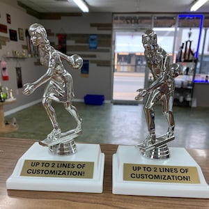 May include: Two silver bowling trophies on white bases with gold plates. The trophies feature a bowler in action, holding a bowling ball. The plates read "UP TO 2 LINES OF CUSTOMIZATION!"