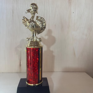 May include: A gold-colored trophy featuring a turkey figure atop a red and gold column. The trophy sits on a black square base. The turkey is wearing a hat. The column has a red, glittery texture.