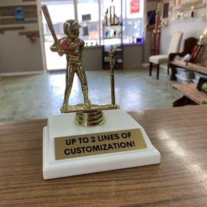 Könnte beinhalten: Goldfarbener Baseballspieler-Pokal mit weißer Basis. Die Basis hat eine goldene Plakette mit dem Text "UP TO 2 LINES OF CUSTOMIZATION!"