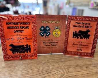 May include: Three orange and brown plaques with black text and images. The left plaque says "Northeast District Livestock Judging Contest 1st Sr. FFA Team". The middle plaque says "Your County Fair" with a 4-H logo and an FFA logo. The right plaque says "Your County Livestock Show Grand Champion 2024".