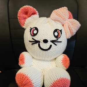 May include: A white crocheted stuffed animal shaped like a cat with pink ears, a pink bow, and pink accents on its paws. The cat has black eyes, a black nose, and a black smiling mouth.