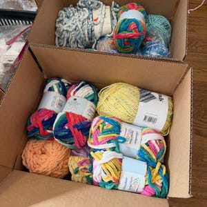 May include: Two cardboard boxes filled with colorful yarn skeins. The yarn is in various colors, including blue, green, yellow, pink, and orange. Some skeins have labels. The yarn appears to be soft and fluffy, suitable for knitting or crocheting projects.
