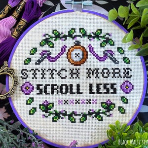 Könnte beinhalten: Ein Kreuzstich-Stickrahmen mit dem Text "STITCH MORE SCROLL LESS" in Schwarz. Das Design umfasst lila und grüne florale Elemente, einen Knopf und eine Schere. Lila Stickgarn und eine Schere sind sichtbar.