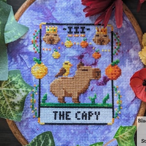 Puede incluir: Aro de bordado de punto de cruz con un fondo morado que presenta un capibara de dibujos animados con un pájaro en su espalda. El diseño está enmarcado en un borde negro con el texto "THE CAPY" en la parte inferior. El aro está rodeado de hojas verdes y flores rojas y amarillas.