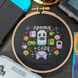 May include: A black embroidery hoop with a white ghost and the words "Spooky Cozy" in colorful pixel art. The embroidery is surrounded by pixelated images of video game controllers, game cartridges, and other gaming-related items.