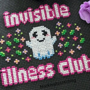 Puede incluir: Dise&ntilde;o de punto de cruz sobre tela negra con un fantasma sonriente con las palabras "Invisible Illness Club" e "Invisible" en rosa y blanco. El fantasma est&aacute; rodeado de flores coloridas.
