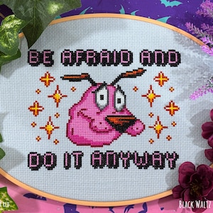 May include: A cross-stitch embroidery hoop featuring Courage the Cowardly Dog with the text "BE AFRAID AND DO IT ANYWAY." The pink dog is surrounded by starbursts. The text and dog are stitched in black and pink thread on a light blue background.