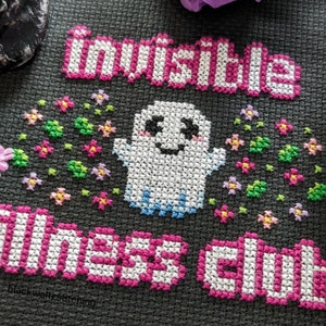 Puede incluir: Dise&ntilde;o de punto de cruz sobre tela gris oscuro con las palabras "invisible illness club" en rosa y blanco. Un fantasma sonriente con los brazos extendidos est&aacute; rodeado de flores coloridas. El texto est&aacute; en un estilo de letras de bloque.