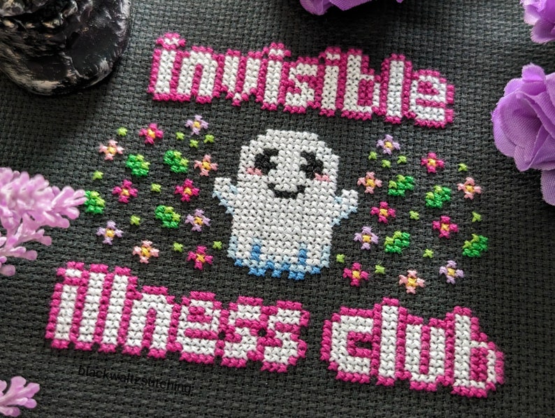 Puede incluir: Dise&ntilde;o de punto de cruz sobre tela gris oscuro con las palabras "invisible illness club" en rosa y blanco. Un fantasma sonriente con los brazos extendidos est&aacute; rodeado de flores coloridas. El texto est&aacute; en un estilo de letras de bloque.