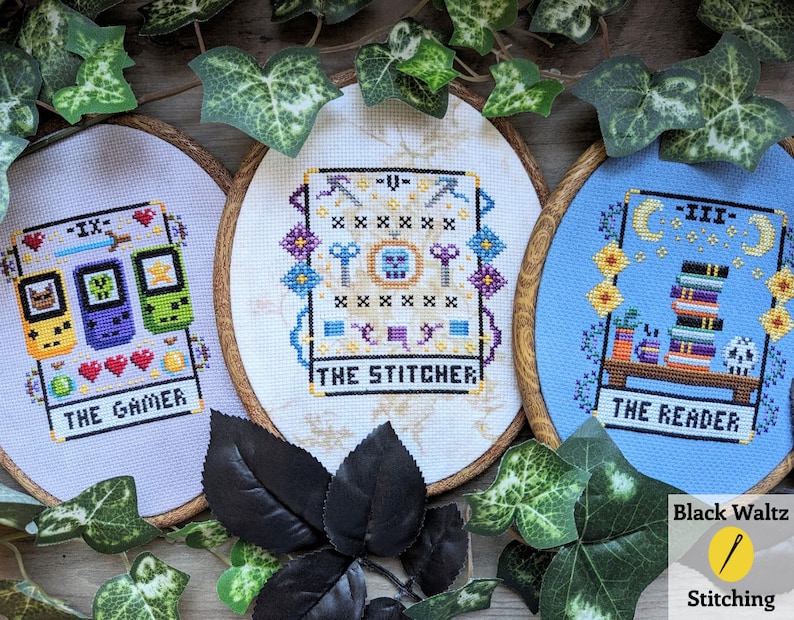 May include: Three embroidery hoops with cross-stitch designs inspired by tarot cards. The first hoop is purple and features a video game controller with the text "The Gamer - IX". The second hoop is white and features a key, a skull, and the text "The Stitcher - V". The third hoop is blue and features a stack of books, a cauldron, and the text "The Reader - III".