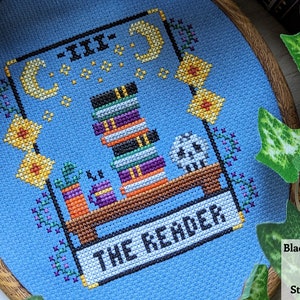 May include: A blue cross-stitch embroidery hoop with a design of a tarot card, The Reader, featuring a stack of books, a crescent moon, and a skull. The card is surrounded by a border of yellow flowers and green vines.