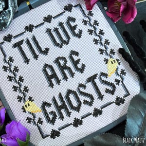 May include: A square cross-stitch embroidery piece featuring the black text 'TIL WE ARE GHOSTS' centered on a light gray fabric. The design is framed by a black border with small heart motifs and two small yellow ghost figures at the corners.