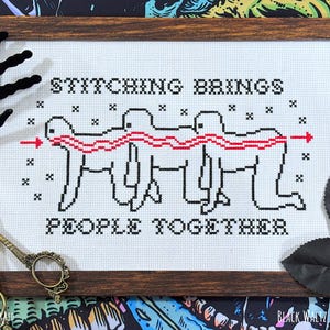 May include: Cross-stitch art in a wooden frame with the text "STITCHING BRINGS PEOPLE TOGETHER." The design features three stick figures connected by a red line, with an arrow. The artwork is on white fabric.