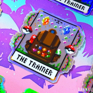 May include: Holographic sticker featuring pixelated art of a brown backpack, gems, and two Pokeballs. The text "THE TRAINER" is displayed on a white banner. The sticker has a colorful, pixelated border and is set against a purple and pink background.