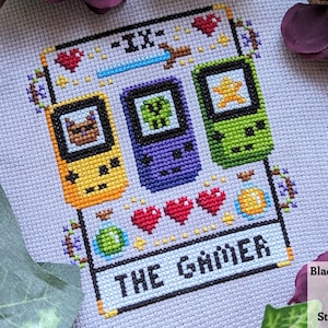 May include: Cross-stitch pattern of a tarot card, The Gamer, with a purple, green, and yellow pixelated game console design. The card is surrounded by hearts, a potion, and a sword.