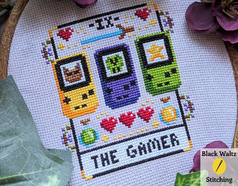 May include: Cross-stitch pattern of a tarot card, The Gamer, with a purple, green, and yellow pixelated game console design. The card is surrounded by hearts, a potion, and a sword.