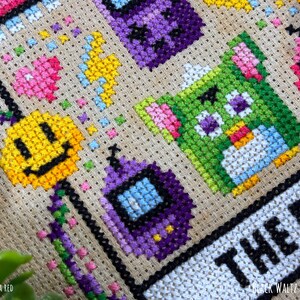 May include: Close-up of a cross-stitch design featuring colorful pixelated characters and shapes. The design includes a yellow smiley face, a green creature, and purple and pink geometric shapes. The text "THE 90'S" is visible.