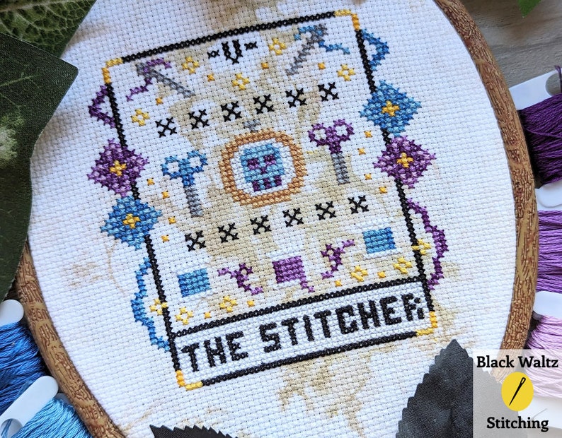May include: Cross-stitch embroidery of a card with the text "The Stitcher" and a design of keys, flowers, and a skull. The card is framed in a wooden hoop.