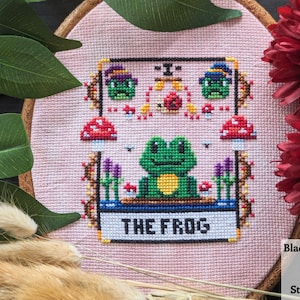 May include: A pink cross-stitch embroidery hoop with a green frog in the center. The frog is surrounded by mushrooms, flowers, and other whimsical details. The text "THE FROGS" is written below the frog.