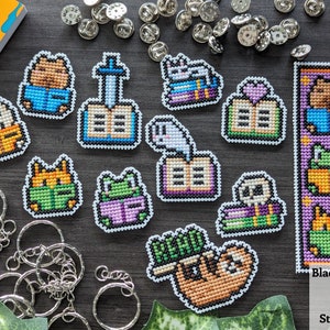 May include: A set of 12 cross-stitch book-themed bookmarks featuring various animals, including a cat, a frog, a sloth, and a bear. The bookmarks are all in different colors and have a white border. The bookmarks are arranged on a dark gray surface with a variety of silver rings and pins.