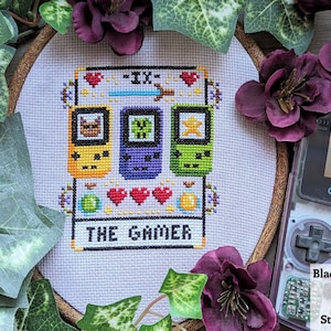 May include: A cross-stitch embroidery hoop with a white background and a pixelated design of three Game Boy Color handhelds. The design includes the text "The Grinder" and a heart symbol.