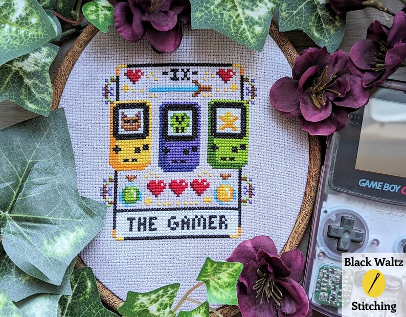May include: A cross-stitch embroidery hoop with a white background and a pixelated design of three Game Boy Color handhelds. The design includes the text "The Grinder" and a heart symbol.