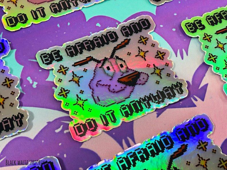 Puede incluir: Pegatina hologr&aacute;fica con Courage el perro cobarde con el texto "BE AFRAID AND DO IT ANYWAY". La pegatina tiene un dise&ntilde;o pixelado con un acabado iridiscente colorido y acentos de estrellas. El fondo es un patr&oacute;n morado y rosa.