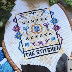 May include: A cross-stitch embroidery hoop with a white fabric background. The design features a skull with a crown, surrounded by keys, axes, and other tools. The text "THE STITCHER" is stitched in black below the design.