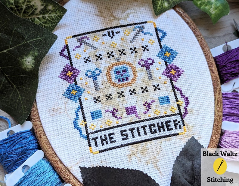 May include: A cross-stitch embroidery hoop with a white fabric background. The design features a skull with a crown, surrounded by keys, axes, and other tools. The text "THE STITCHER" is stitched in black below the design.