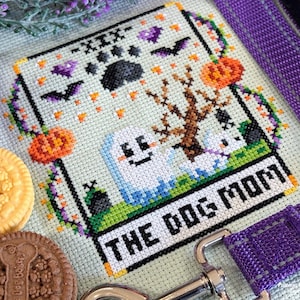 Op de afbeelding: Kruissteekontwerp met een Halloween-thema, met geesten, pompoenen en vleermuizen. De tekst "THE DOG MOM" is prominent aanwezig. Inclusief hondenkoekjes en een paarse hondenriem.