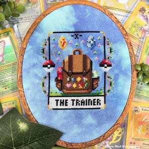May include: Cross-stitch embroidery in a wooden hoop featuring a brown backpack with colorful embellishments, two Poké Balls, and the text "THE TRAINER" on a light blue background. Surrounded by Pokémon trading cards.