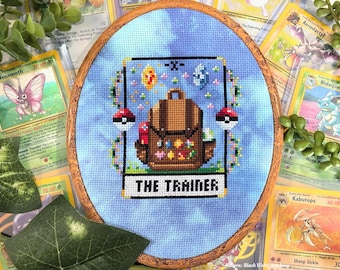 May include: Cross-stitch embroidery in a wooden hoop featuring a brown backpack with colorful embellishments, two Poké Balls, and the text "THE TRAINER" on a light blue background. Surrounded by Pokémon trading cards.