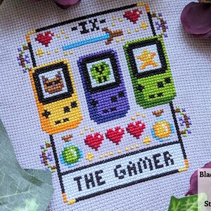 May include: Cross-stitch pattern of a tarot card, The Gamer, with a purple, green, and yellow pixelated game console design. The card is surrounded by hearts, a potion, and a sword.