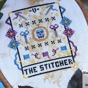 May include: Cross-stitch embroidery hoop with a white fabric background. The design features a black border with the text "THE STITCHER" in black lettering. The center of the design features a colorful pattern with a skull and crossbones in the center.
