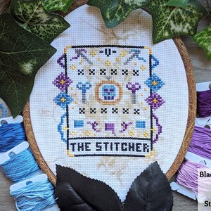 May include: A white embroidery hoop with a cross-stitch design featuring a skull, keys, and scissors. The design is surrounded by a colorful border and the text "The Stitcher" is below the design.